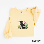 Butter crewneck sweatshirt laid flat on white, embroidered with a sitting black Labrador silhouette among yellow bell flowers, a white tulip and pink daisies, with a pink butterfly