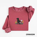 Crimson crewneck sweatshirt laid flat on white, embroidered with a sitting black Labrador silhouette among yellow bell flowers, a white tulip and pink daisies, with a pink butterfly