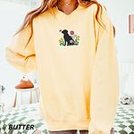 Woman in an oversized Butter crewneck sweatshirt embroidered with a sitting black Labrador silhouette among yellow bell flowers, a white tulip and pink daisies, with a pink butterfly