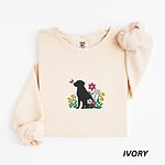 Ivory crewneck sweatshirt laid flat on white, embroidered with a sitting black Labrador silhouette among yellow bell flowers, a white tulip and pink daisies, with a pink butterfly