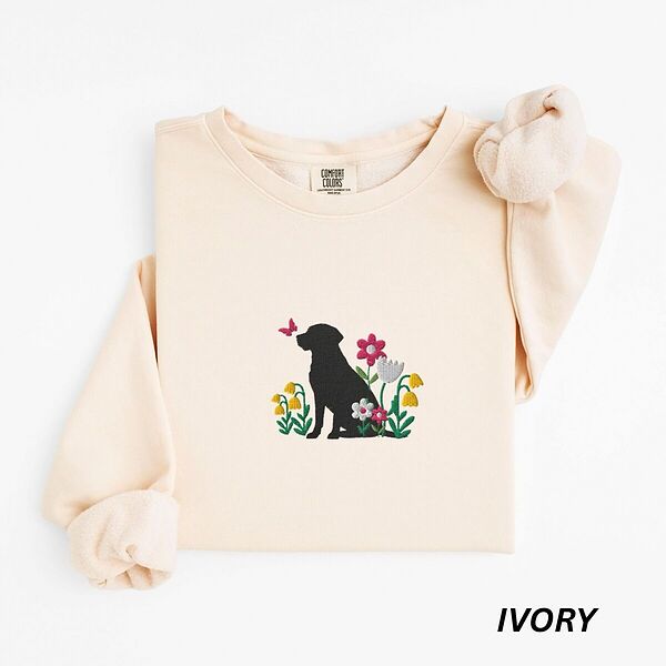 Ivory crewneck sweatshirt laid flat on white, embroidered with a sitting black Labrador silhouette among yellow bell flowers, a white tulip and pink daisies, with a pink butterfly