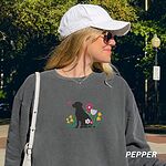 Woman in a white cap walking outdoors in a Pepper crewneck sweatshirt embroidered with a sitting black Labrador silhouette among yellow bell flowers, a white tulip and pink daisies, with a pink butterfly