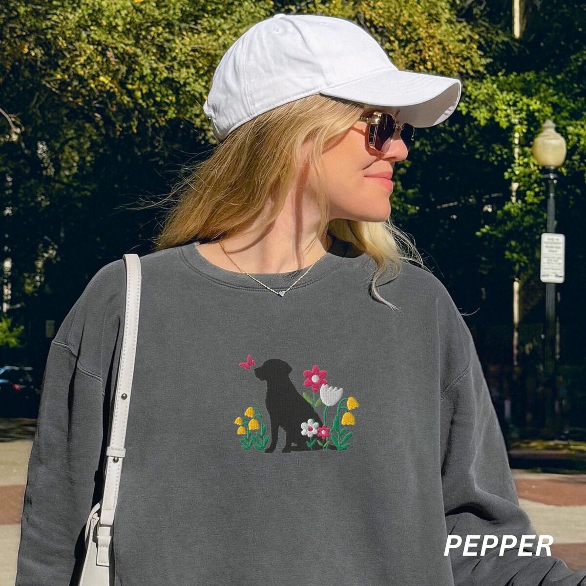 Floral Black Lab Sweatshirt – Pepper Woman in a white cap walking outdoors in a Pepper crewneck sweatshirt embroidered with a sitting black Labrador silhouette among yellow bell flowers, a white tulip and pink daisies, with a pink butterfly