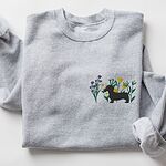 Grey crewneck sweatshirt laid flat on white, embroidered with a black dachshund silhouette standing among purple, yellow and white wildflowers on the left chest