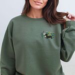 Woman in a Military Green crewneck sweatshirt embroidered with a black dachshund silhouette standing among purple, yellow and white wildflowers on the left chest