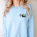 Woman in a Light Blue crewneck sweatshirt embroidered with a black dachshund silhouette standing among purple, yellow and white wildflowers on the left chest