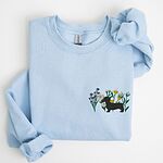 Light Blue crewneck sweatshirt laid flat on white, embroidered with a black dachshund silhouette standing among purple, yellow and white wildflowers on the left chest