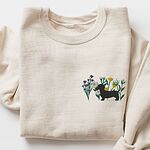 Sand crewneck sweatshirt laid flat on white, embroidered with a black dachshund silhouette standing among purple, yellow and white wildflowers on the left chest