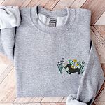 Sport Grey crewneck sweatshirt laid flat on herringbone wood, embroidered with a black dachshund silhouette standing among purple, yellow and white wildflowers on the left chest