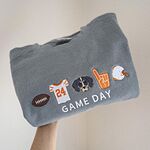 Grey Comfort Colors crewneck sweatshirt held up folded, embroidered with a football, an orange and white jersey with the number 24, a black and tan hound face, an orange foam finger and a white helmet above the words GAME DAY