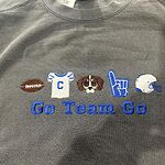 Pepper Comfort Colors crewneck sweatshirt embroidered with a football, a white jersey with a blue letter C, a brown and white beagle face, a royal blue foam finger and a white helmet above the words Go Team Go in royal blue