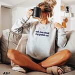 Woman sitting on a couch with a black mug in an Ash crewneck sweatshirt, with the words ask me about my foster cat embroidered in cursive across the chest in navy thread
