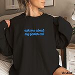 Woman in a Black crewneck sweatshirt in a bedroom, with the words ask me about my foster cat embroidered in cursive across the chest in bright blue thread