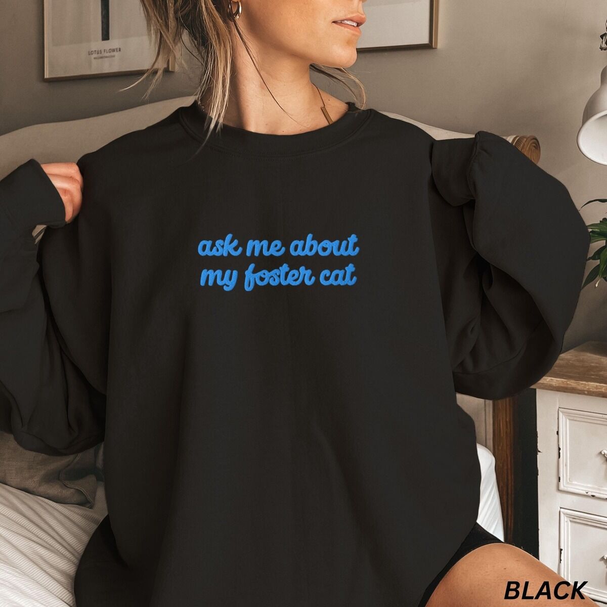 Foster Cat Sweatshirt – Black Woman in a Black crewneck sweatshirt in a bedroom, with the words ask me about my foster cat embroidered in cursive across the chest in bright blue thread
