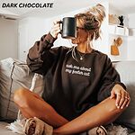 Woman sitting on a couch with a black mug in a Dark Chocolate crewneck sweatshirt, with the words ask me about my foster cat embroidered in cursive across the chest in white thread