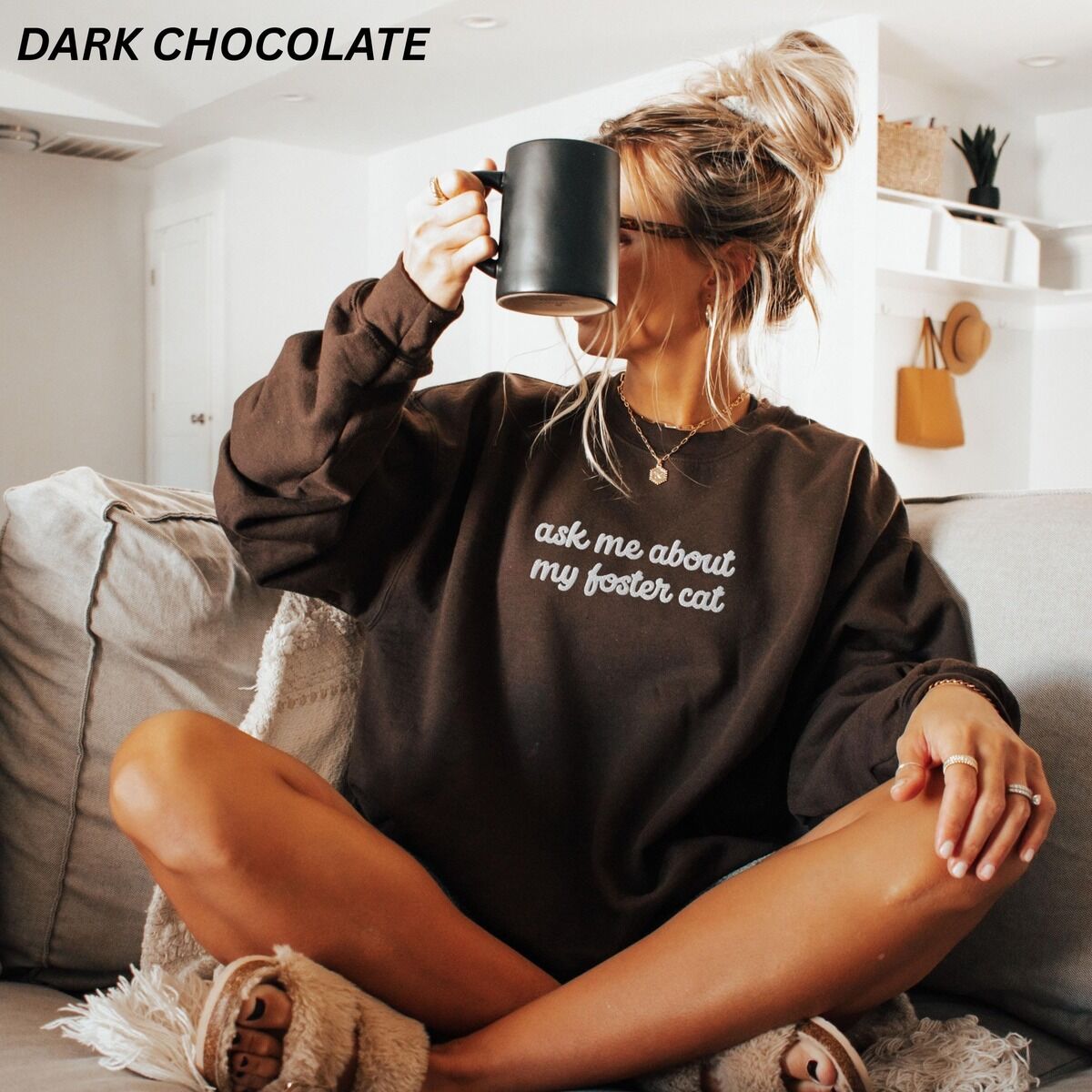 Foster Cat Sweatshirt – Dark Chocolate Woman sitting on a couch with a black mug in a Dark Chocolate crewneck sweatshirt, with the words ask me about my foster cat embroidered in cursive across the chest in white thread