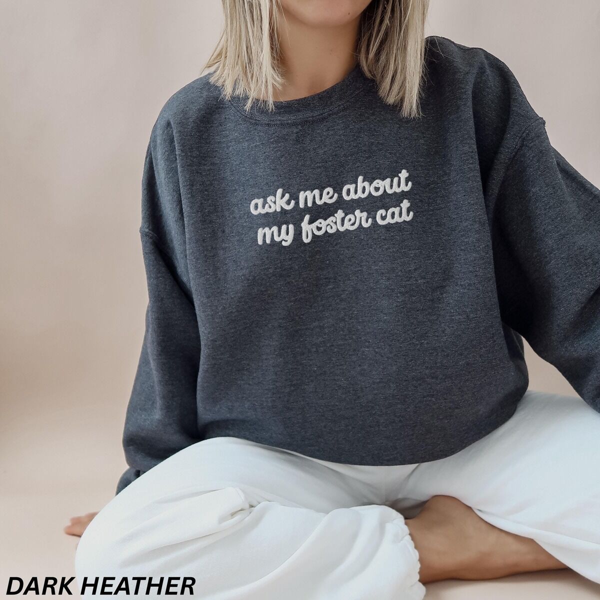 Foster Cat Sweatshirt – Dark Heather Woman sitting cross-legged in a Dark Heather crewneck sweatshirt, with the words ask me about my foster cat embroidered in cursive across the chest in white thread