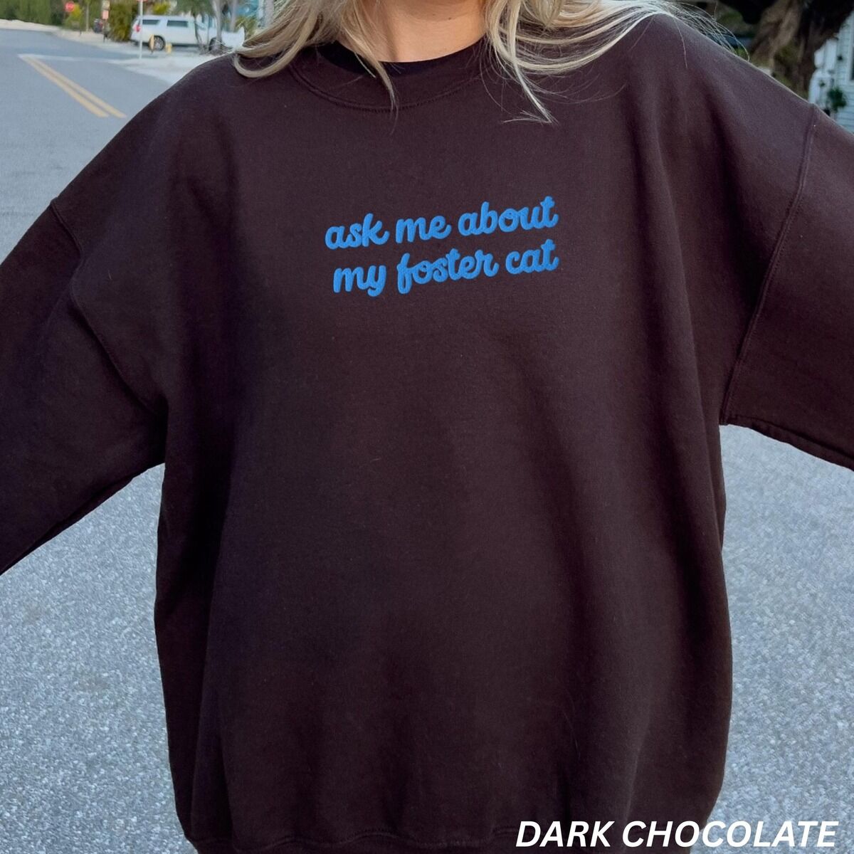 Foster Cat Sweatshirt Blue Thread Woman standing on a street in a Dark Chocolate crewneck sweatshirt, with the words ask me about my foster cat embroidered in cursive across the chest in bright blue thread