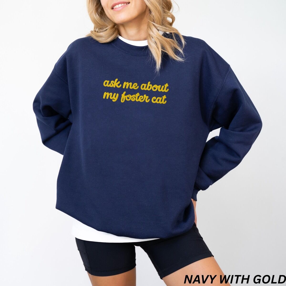 Foster Cat Sweatshirt Gold Thread Woman in a Navy crewneck sweatshirt with the words ask me about my foster cat embroidered in cursive across the chest in gold thread