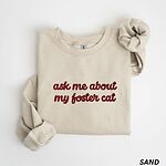 Sand crewneck sweatshirt folded on a white background, with the words ask me about my foster cat embroidered in cursive across the chest in red thread