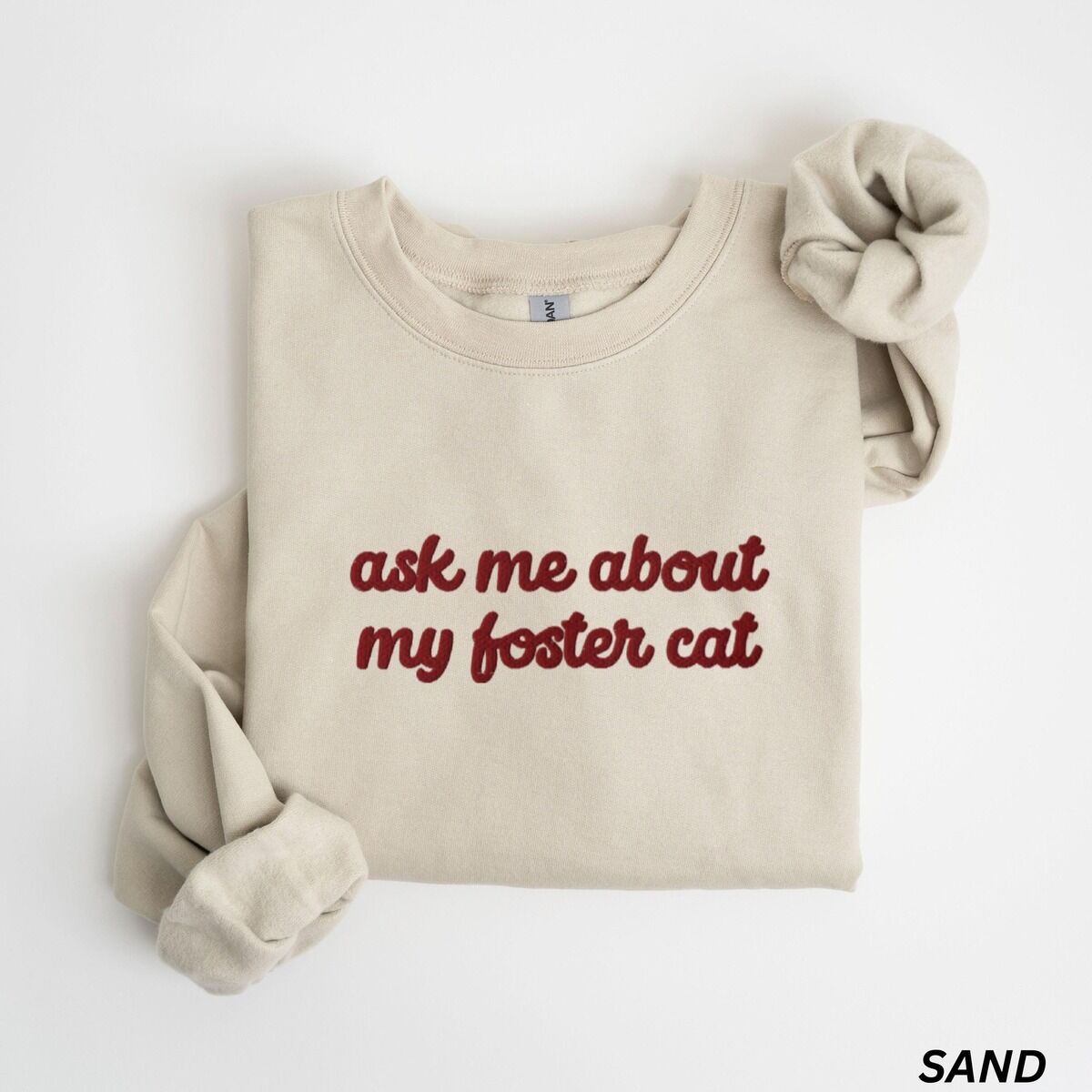Foster Cat Sweatshirt Red Thread Sand crewneck sweatshirt folded on a white background, with the words ask me about my foster cat embroidered in cursive across the chest in red thread