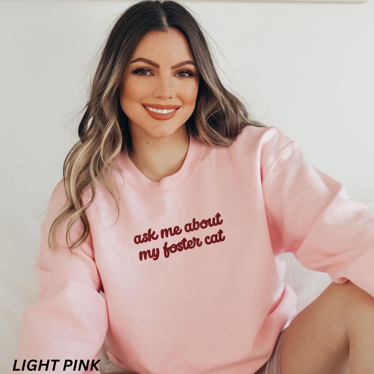 Foster Cat Sweatshirt Light Pink Smiling woman in a Light Pink crewneck sweatshirt with the words ask me about my foster cat embroidered in cursive across the chest in deep red thread