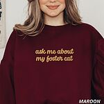 Woman in a Maroon crewneck sweatshirt with the words ask me about my foster cat embroidered in cursive across the chest in gold thread