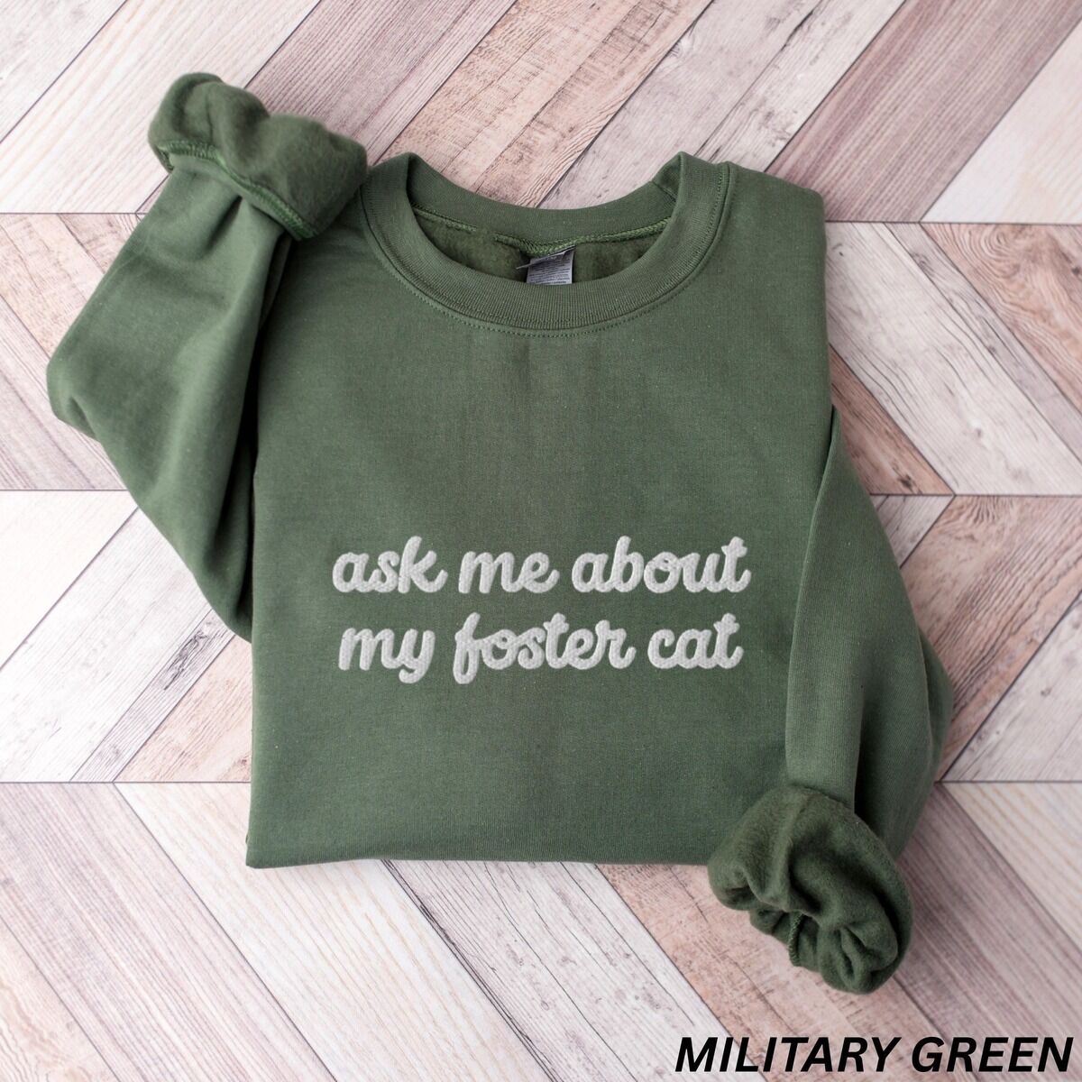 Foster Cat Sweatshirt – Military Green Military Green crewneck sweatshirt folded on a herringbone wood floor, with the words ask me about my foster cat embroidered in cursive across the chest in white thread