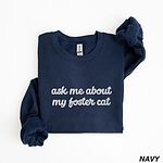 Navy crewneck sweatshirt folded on a white background, with the words ask me about my foster cat embroidered in cursive across the chest in white thread