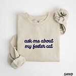 Sand foster cat sweatshirt folded on a white background, with the words ask me about my foster cat embroidered in cursive across the chest in navy thread