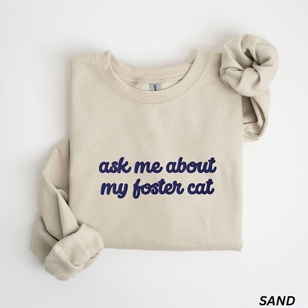 Sand foster cat sweatshirt folded on a white background, with the words ask me about my foster cat embroidered in cursive across the chest in navy thread