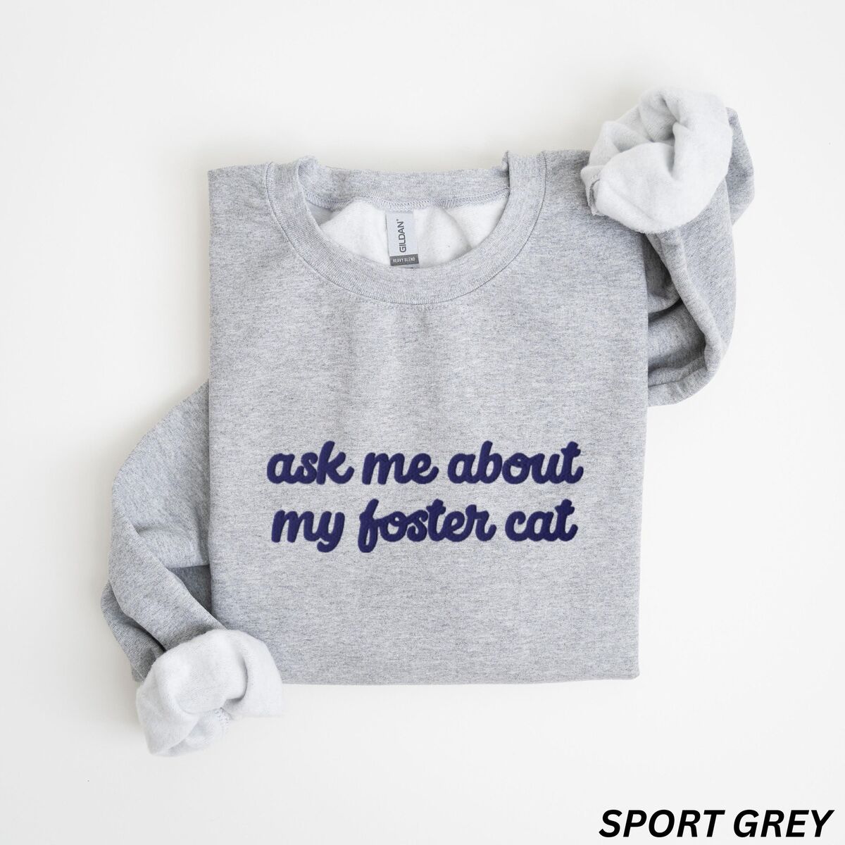 Foster Cat Sweatshirt Sport Grey Sport Grey crewneck sweatshirt folded on a white background, with the words ask me about my foster cat embroidered in cursive across the chest in navy thread