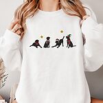 Woman holding the collar of a white crewneck sweatshirt embroidered with four black Labrador silhouettes in a bow, a candy-cane scarf, a Santa hat and an elf hat under gold stars