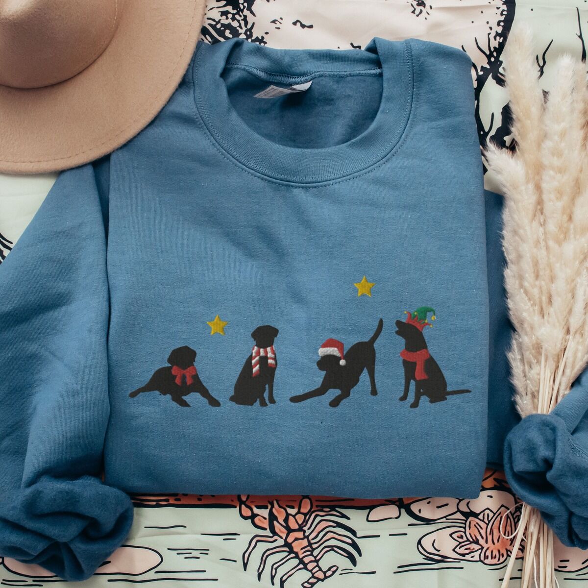 Four Black Labs Christmas Sweatshirt – Indigo Blue Indigo Blue crewneck sweatshirt laid flat beside a hat and pampas grass, embroidered with four black Labrador silhouettes in a bow, a candy-cane scarf, a Santa hat and an elf hat under gold stars