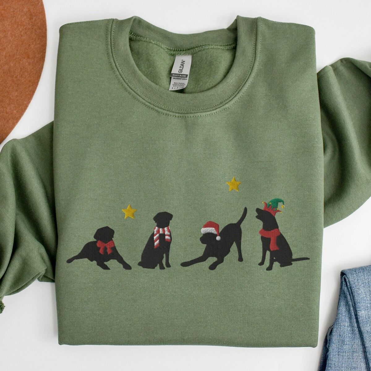 Four Black Labs Christmas Sweatshirt – Military Green Military Green crewneck sweatshirt laid flat beside a rolled pair of jeans, embroidered with four black Labrador silhouettes in a bow, a candy-cane scarf, a Santa hat and an elf hat under gold stars