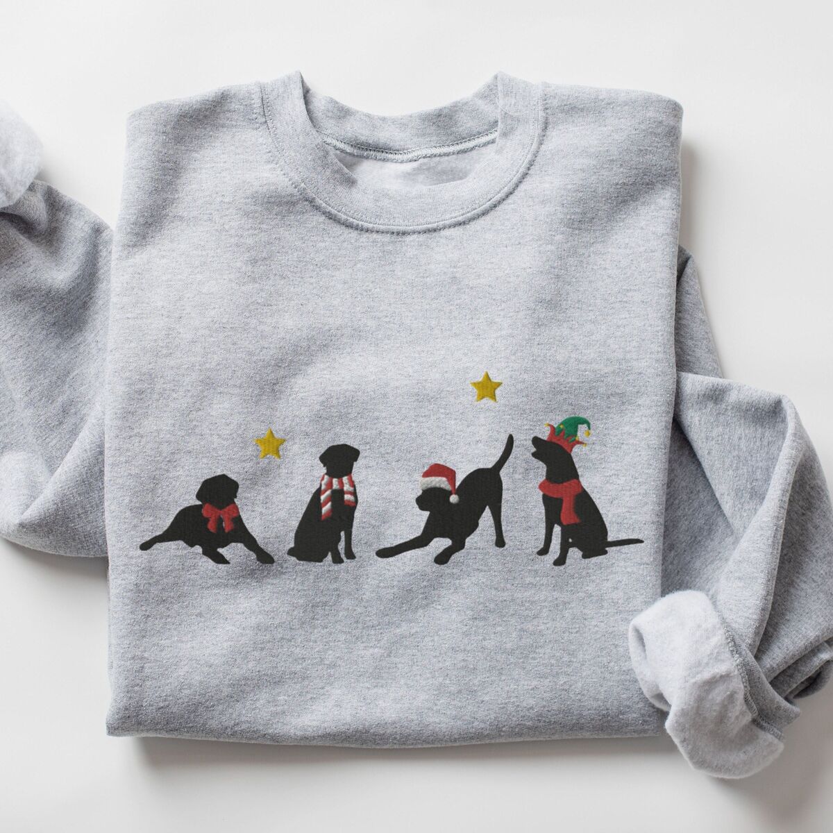Four Black Labs Christmas Sweatshirt - Sport Grey Sport Grey crewneck sweatshirt laid flat with folded sleeves, embroidered with four black Labrador silhouettes in a bow, a candy-cane scarf, a Santa hat and an elf hat under gold stars