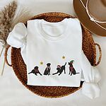 White crewneck sweatshirt folded in a wicker tray beside a hat, embroidered with four black Labrador silhouettes in a bow, a candy-cane scarf, a Santa hat and an elf hat under gold stars