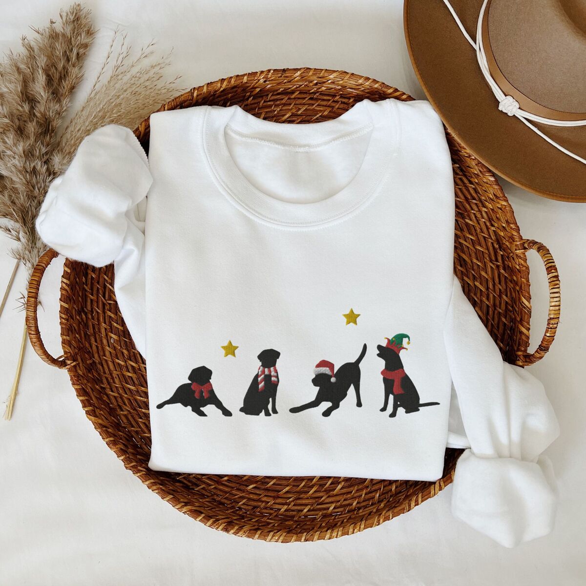 Four Black Labs Christmas Sweatshirt - White White crewneck sweatshirt folded in a wicker tray beside a hat, embroidered with four black Labrador silhouettes in a bow, a candy-cane scarf, a Santa hat and an elf hat under gold stars