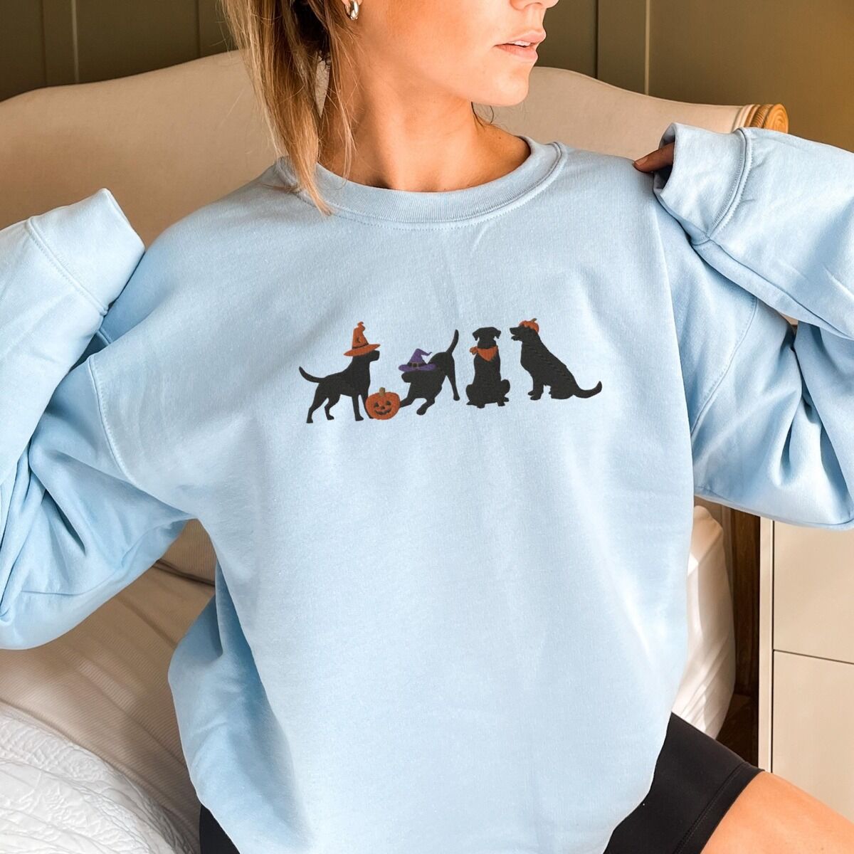 Four Black Labs Halloween Sweatshirt - worn Woman sitting in a light blue crewneck sweatshirt embroidered with four black lab silhouettes in a row, two in witch hats, one in a bandana and one with a tiny pumpkin hat, beside a jack-o'-lantern