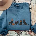 Indigo Blue crewneck sweatshirt laid flat with a brown hat and pampas grass, embroidered with four black lab silhouettes in a row, two in witch hats, one in a bandana and one with a tiny pumpkin hat, beside a jack-o'-lantern