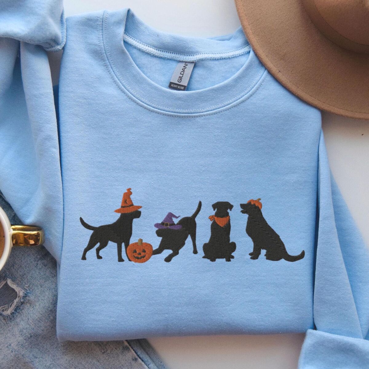 Four Black Labs Halloween Sweatshirt - Light Blue Light Blue crewneck sweatshirt laid flat beside jeans and a brown hat, embroidered with four black lab silhouettes in a row, two in witch hats, one in a bandana and one with a tiny pumpkin hat, beside a jack-o'-lantern
