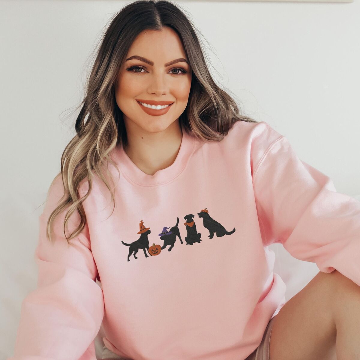 Four Black Labs Halloween Sweatshirt - Light Pink Smiling woman sitting in a Light Pink crewneck sweatshirt embroidered with four black lab silhouettes in a row, two in witch hats, one in a bandana and one with a tiny pumpkin hat, beside a jack-o'-lantern