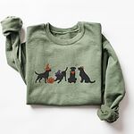 Military Green crewneck sweatshirt laid flat on a white background, embroidered with four black lab silhouettes in a row, two in witch hats, one in a bandana and one with a tiny pumpkin hat, beside a jack-o'-lantern