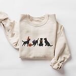 Sand crewneck sweatshirt laid flat on a white background, embroidered with four black lab silhouettes in a row, two in witch hats, one in a bandana and one with a tiny pumpkin hat, beside a jack-o'-lantern
