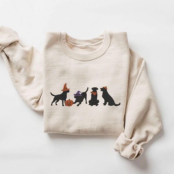 Sand crewneck sweatshirt laid flat on a white background, embroidered with four black lab silhouettes in a row, two in witch hats, one in a bandana and one with a tiny pumpkin hat, beside a jack-o'-lantern