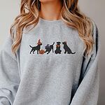 Woman with long blonde hair in a Sport Grey crewneck sweatshirt embroidered with four black lab silhouettes in a row, two in witch hats, one in a bandana and one with a tiny pumpkin hat, beside a jack-o'-lantern