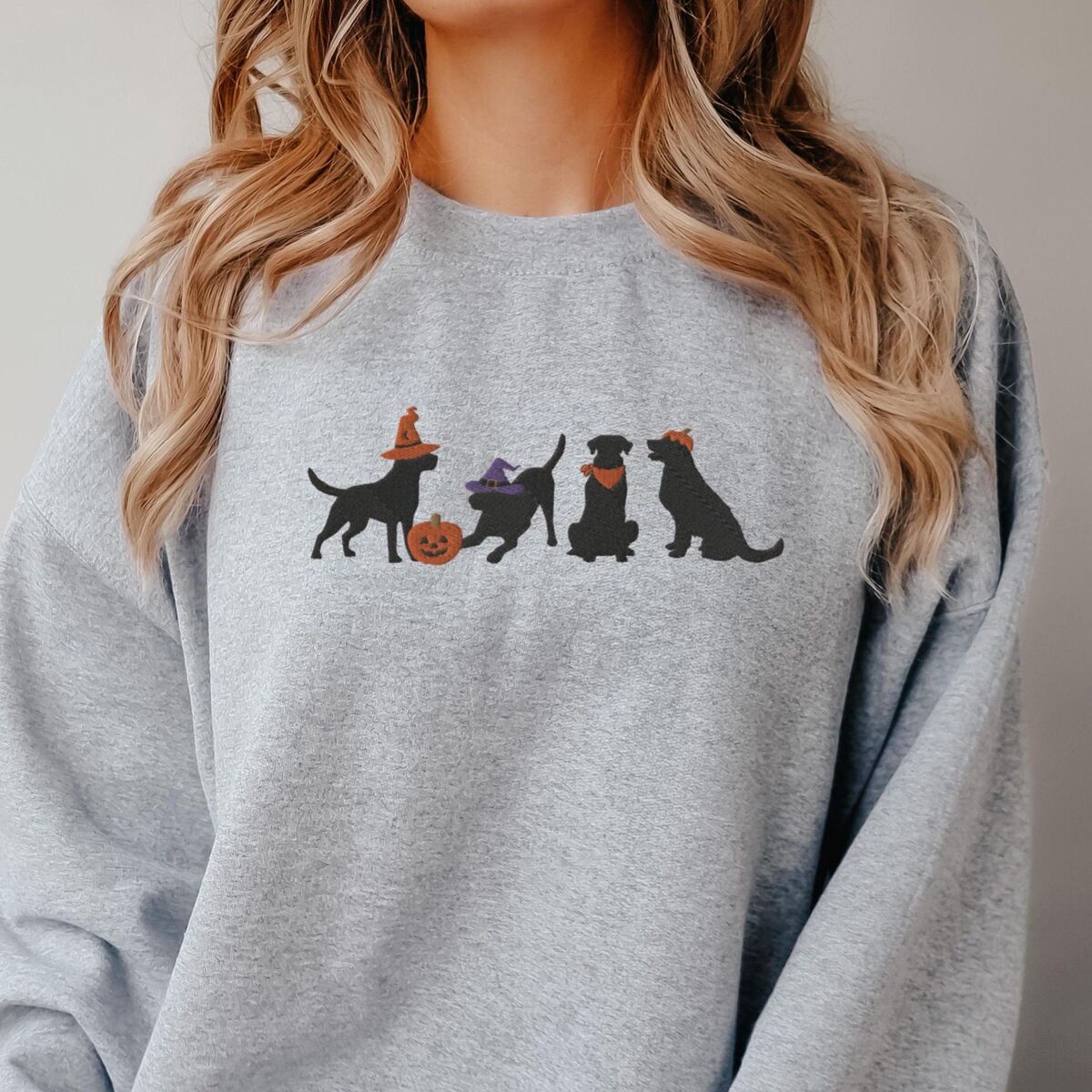 Four Black Labs Halloween Sweatshirt - Sport Grey Woman with long blonde hair in a Sport Grey crewneck sweatshirt embroidered with four black lab silhouettes in a row, two in witch hats, one in a bandana and one with a tiny pumpkin hat, beside a jack-o'-lantern