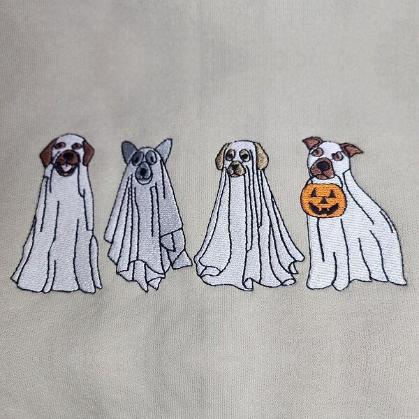 Close-up of the embroidery: four dogs in ghost sheets with brown, gray and golden ears, the last one holding an orange jack-o'-lantern bucket, stitched on a Sand sweatshirt