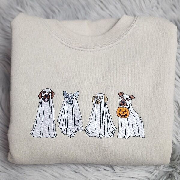Sand crewneck sweatshirt folded on a gray faux fur rug, embroidered with four dogs in white ghost sheets standing in a row, the last one holding a jack-o'-lantern bucket