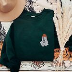 Forest Green crewneck sweatshirt laid flat on a printed backdrop with a felt hat and pampas grass, embroidered with a little ghost in a rust knit beanie hugging a grey French bulldog on the left chest